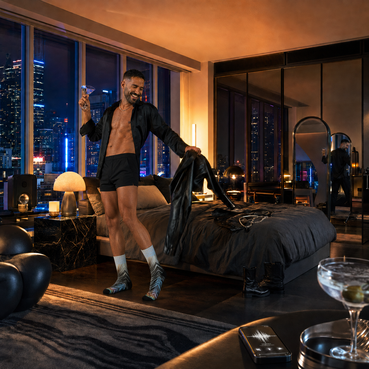 Dani dances in Palm of Pride socks and short black shorts while holding a martini and leather jacket in a luxurious penthouse bedroom.