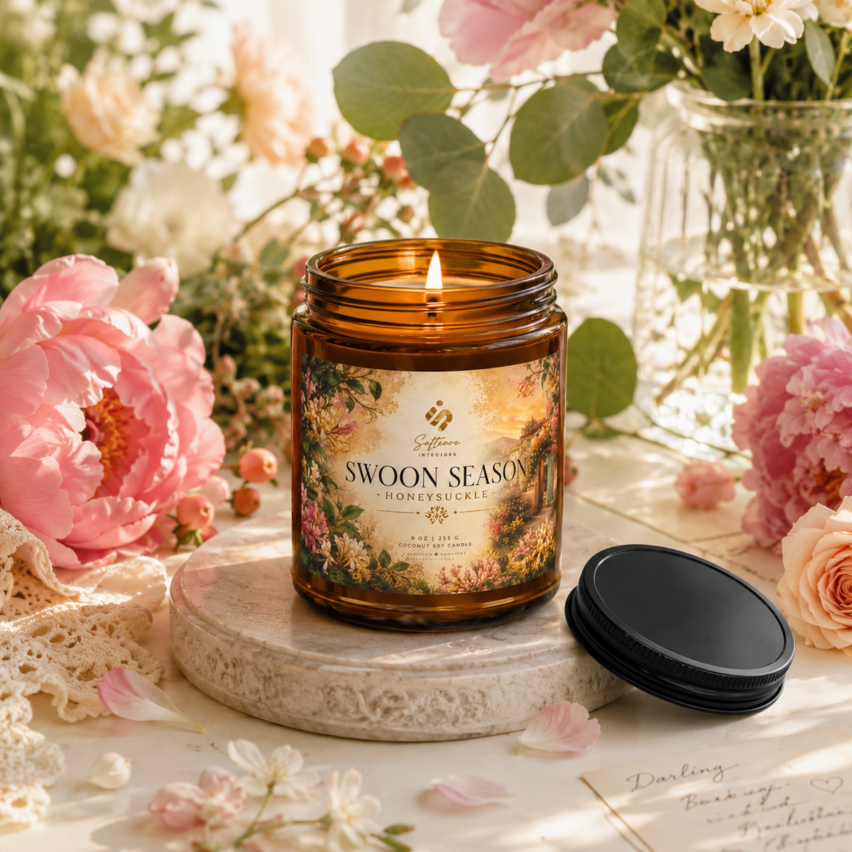 A lit amber-jar Swoon Season candle on a stone tray surrounded by pink flowers.