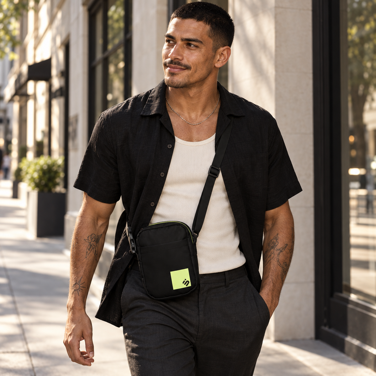 A person in a black overshirt and trousers wearing the Off Record black crossbody bag on a sunlit sidewalk.