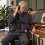 A person listening through headphones in a record shop while wearing the purple Velvet Detour crossbody bag.
