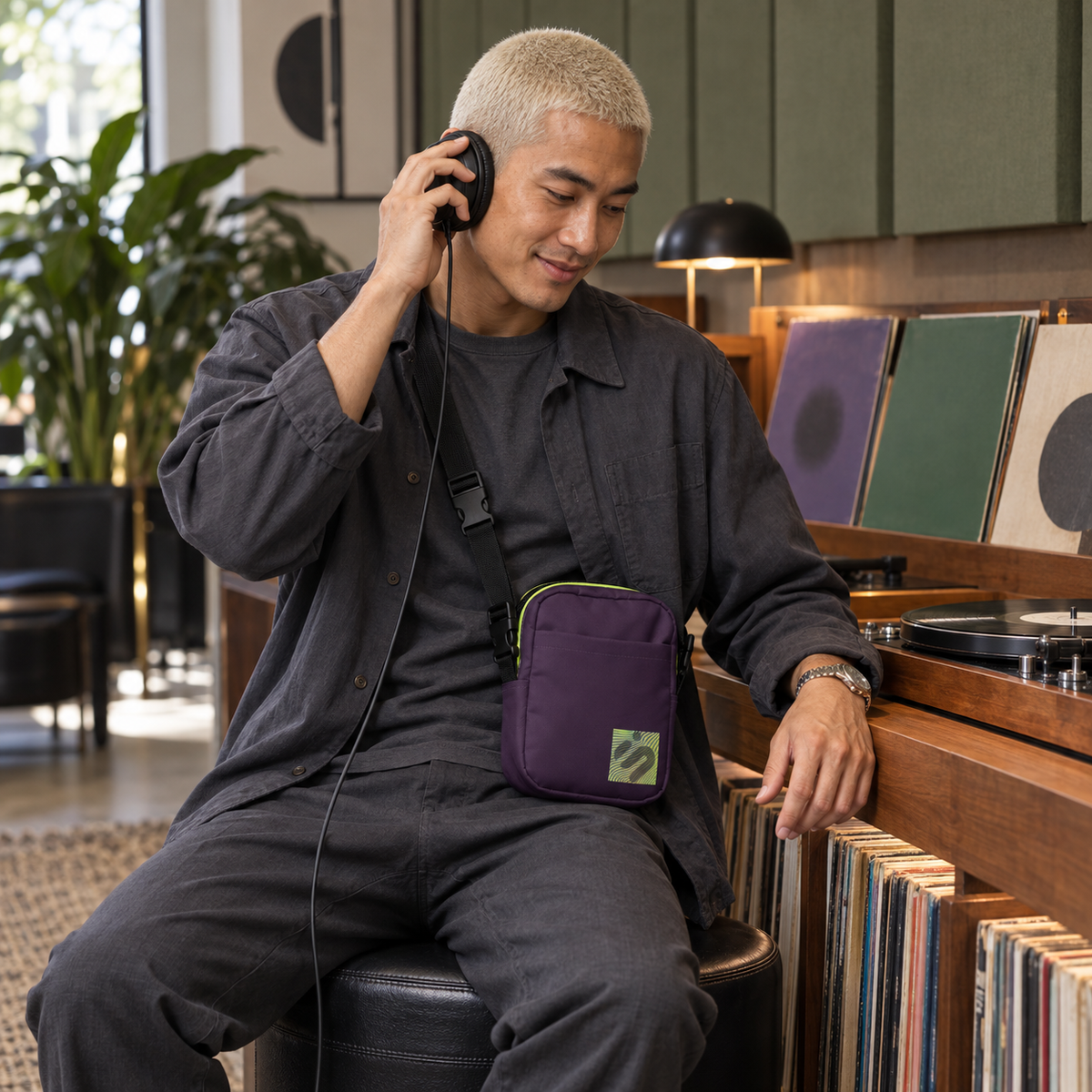 A person listening through headphones in a record shop while wearing the purple Velvet Detour crossbody bag.
